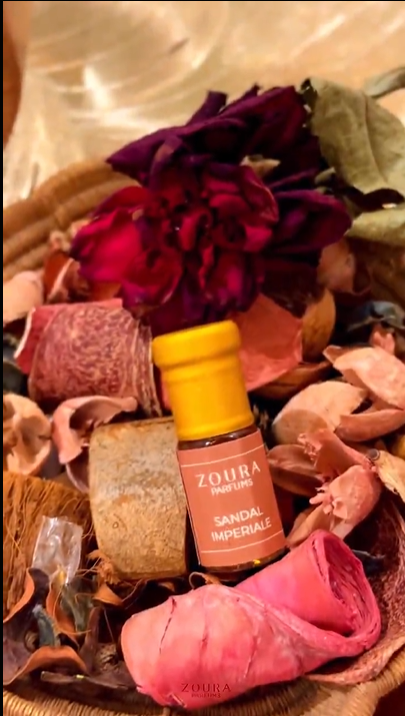 Woman speaking on camera reviewing Zoura Parfums attar, seated indoors with plant and curtain backdrop.