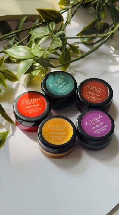 Five Zoura Parfums 8g solid perfume jars in colorful lids — including Red Affair, Melon, Sunshine and Lavish Oud — styled with greenery.