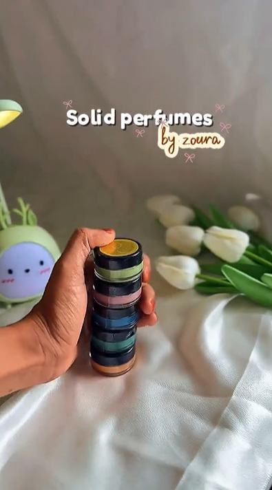 Hand holding an 8g Zoura Parfums solid perfume jar amid tulips and decor, "Solid Perfume by Zoura" showcase.