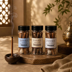 Zoura bakhoor sticks trio – Oud Eclat, Zahra Al Haya, and Oud Noir jars with a burning bakhoor stick in a holder, styled with Moroccan lattice window