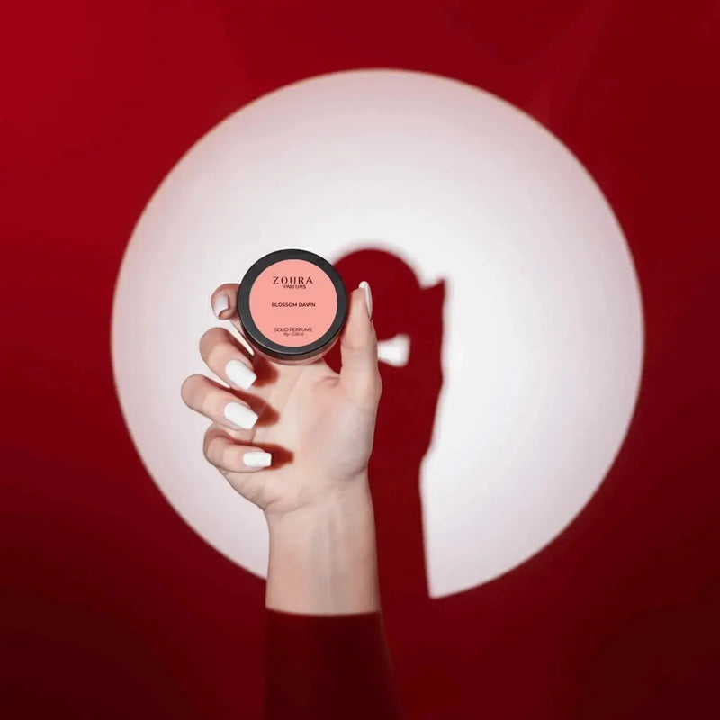 Hand holding Zoura Blossom Dawn Luxury Solid Perfume jar in spotlight against red background