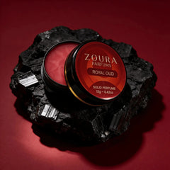 Open jar of Zoura Royal Oud Unisex Solid Perfume resting on black charcoal stone, red backdrop