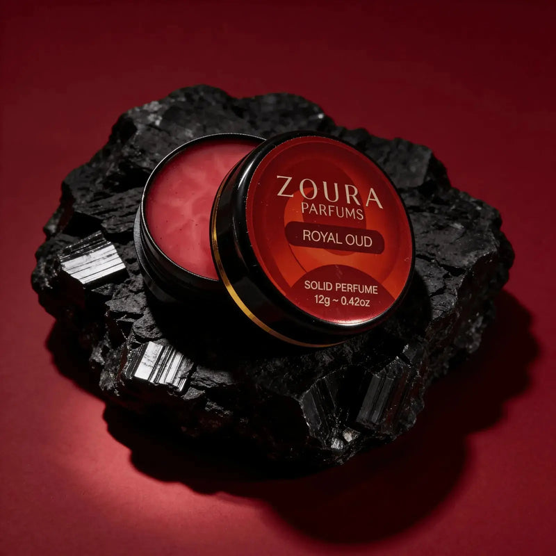 Open jar of Zoura Royal Oud Unisex Solid Perfume resting on black charcoal stone, red backdrop