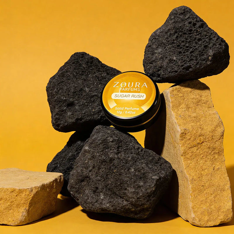 Zoura Sugar Rush Unisex Solid Perfume jar nestled among stacked stones on yellow backdrop